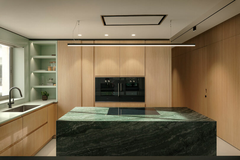 Modern kitchen with light wood cabinets, a large green marble island, built-in ovens, and a rectangular ceiling light fixture.