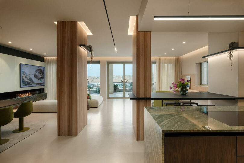 Modern open-plan living area with wooden accents, featuring a kitchen, a dining table with green chairs, a wall-mounted TV, and large windows overlooking a balcony with a view.
