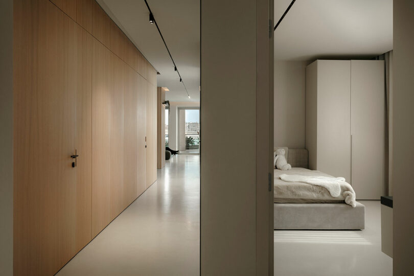 Minimalist interior with a hallway featuring wooden paneling. A door opens to a bedroom with a bed, white linens, and a wardrobe. A view outside is visible at the end of the hall.