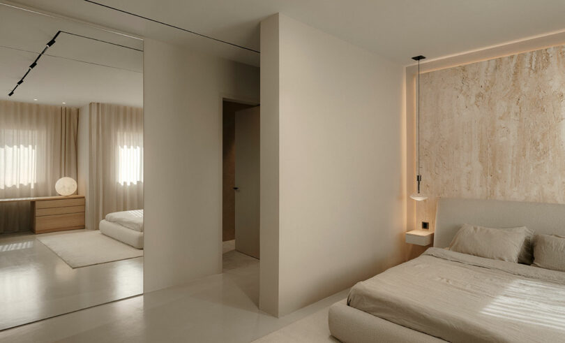Minimalist bedroom with beige tones, featuring a bed with a textured accent wall, a hanging light, and a mirrored closet. Natural light enters through the window with sheer curtains.