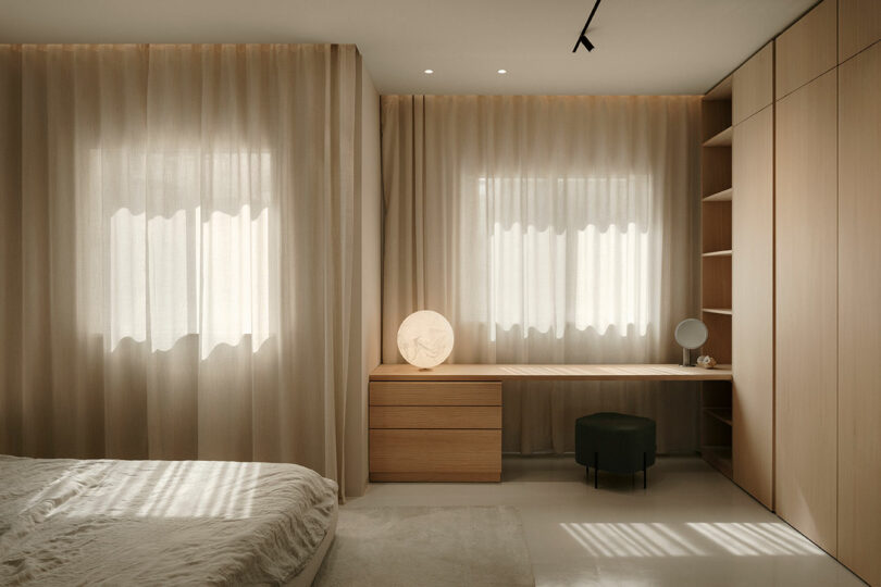 Minimalist bedroom with beige curtains, a wooden desk, and a dresser. A round lamp and circular mirror are on the desk. Neutral tones and natural light create a serene atmosphere.