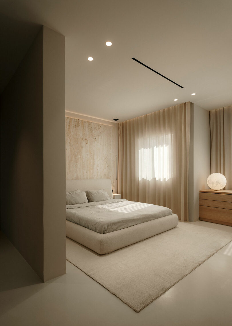 Minimalist bedroom with a low bed, light bedding, a textured wall, sheer curtains, and a wooden dresser. A spherical lamp sits on the dresser, and warm lighting illuminates the room.
