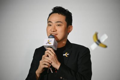 A man stands in front of a banana duct-taped to a wall holding a microphone. 