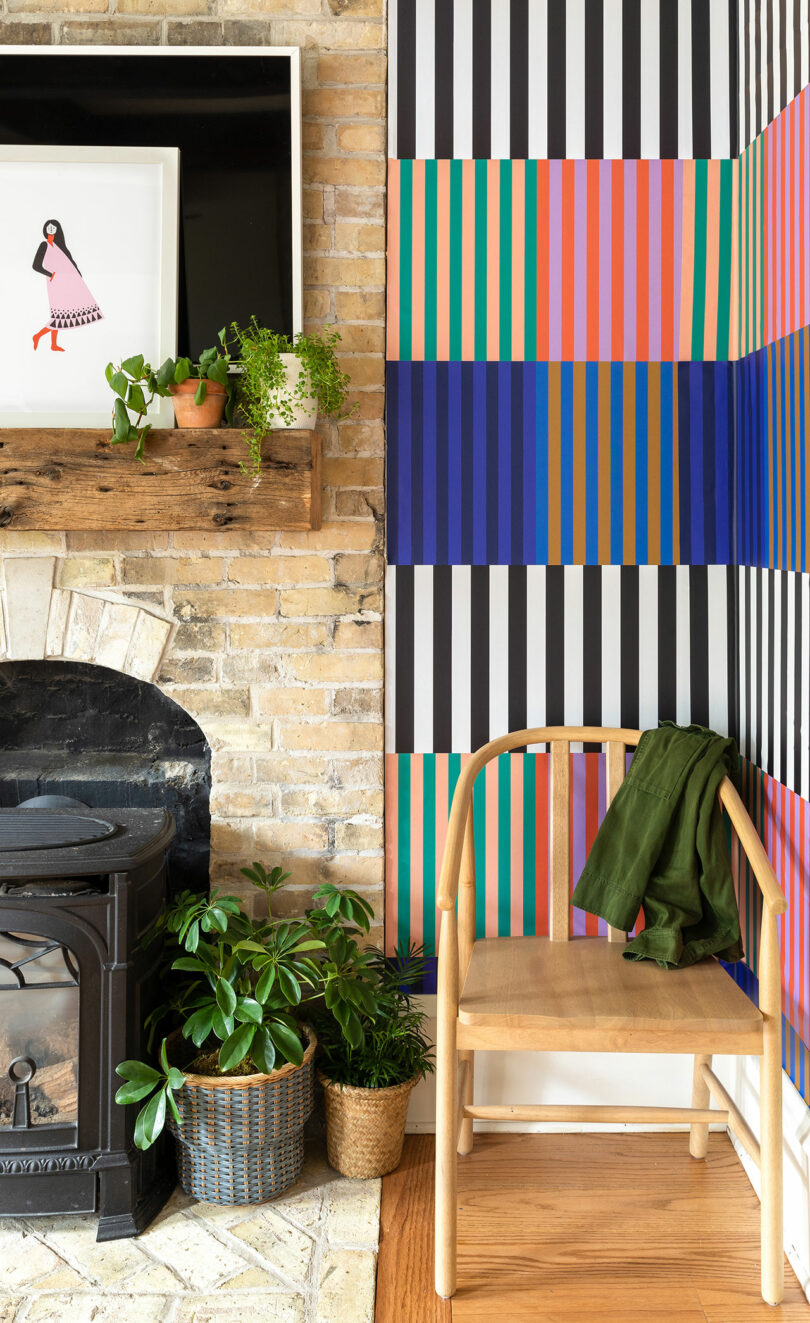 A wooden chair with a green jacket sits beside a colorful striped wall designed by Elizabeth Rees, potted plants, a brick fireplace, and a mantle with framed art and more plants.