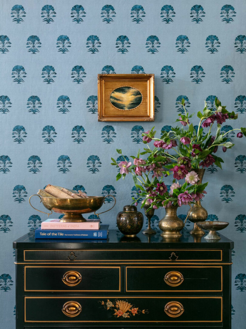 A black chest with floral detailing, designed by Elizabeth Rees, holds brass bowls, a vase with purple flowers, and stacked books, set against blue wallpaper and a small framed painting above.
