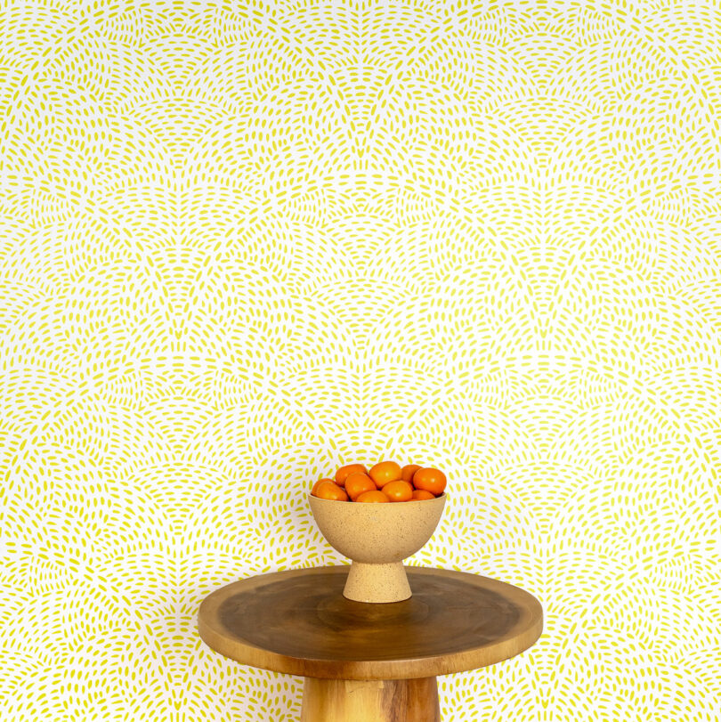A ceramic bowl filled with cherry tomatoes sits on a round wooden table against a patterned yellow and white Elizabeth Rees background.
