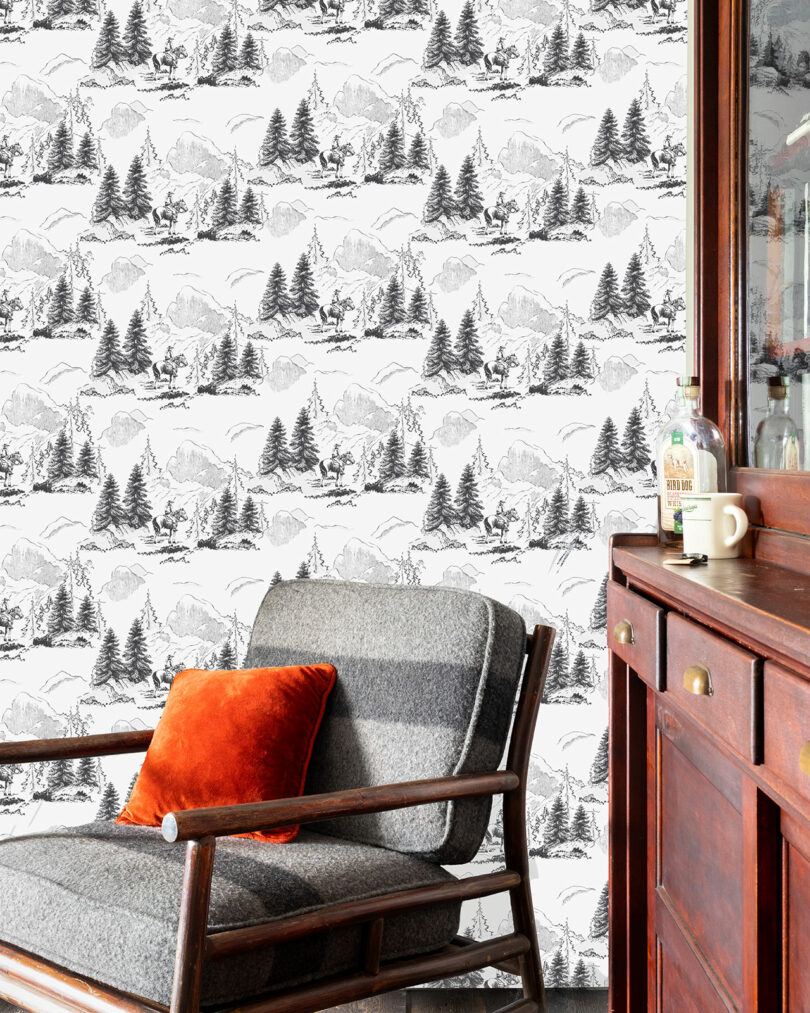 A gray and wood armchair with an orange pillow sits next to a wooden cabinet; the wall features black and white landscape-patterned wallpaper by Elizabeth Rees.