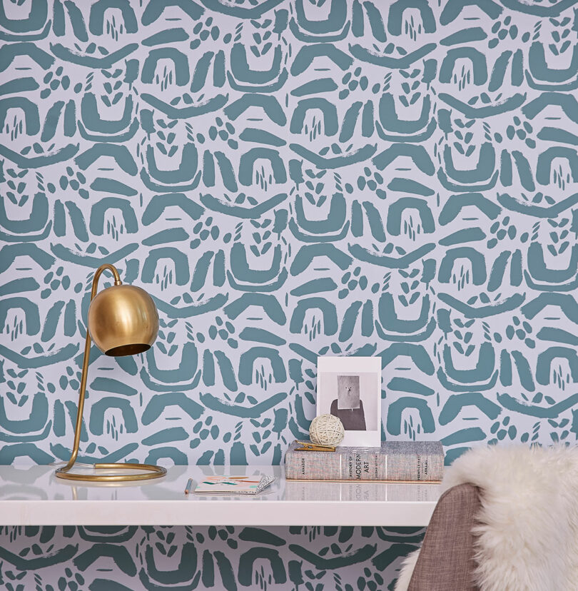 A minimalist desk with a gold lamp, stacked books, framed art, and a fuzzy chair sits against blue and white abstract patterned wallpaper by Elizabeth Rees.