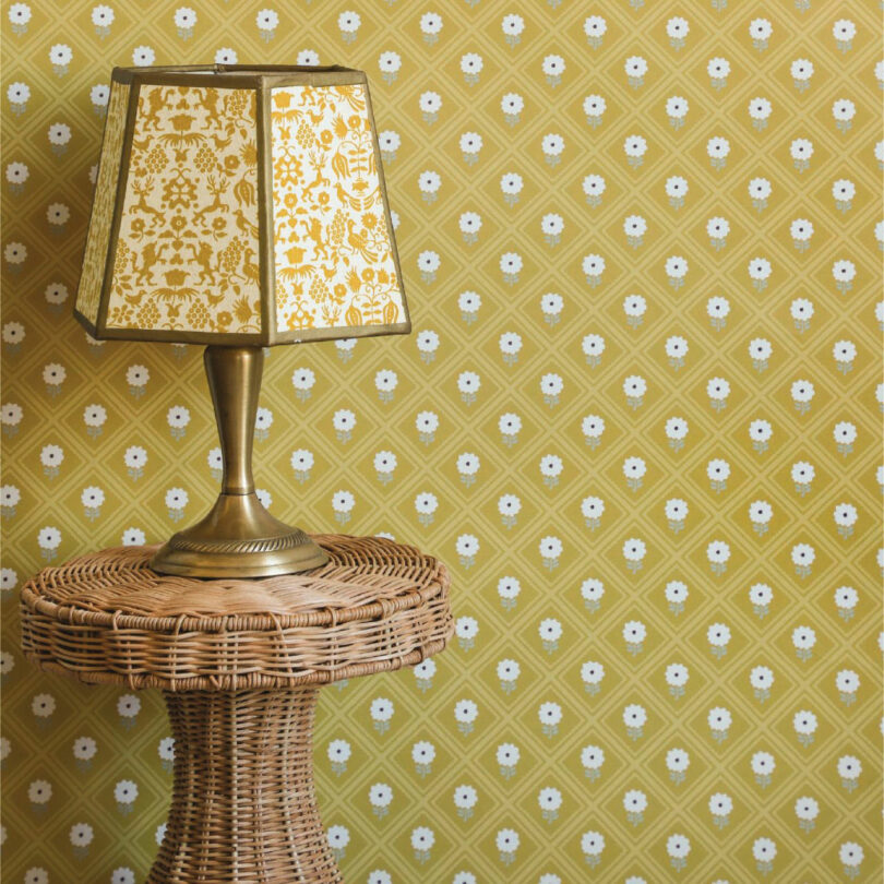 A wicker side table with a brass lamp featuring a patterned shade from Elizabeth Rees sits in front of yellow floral-patterned wallpaper.