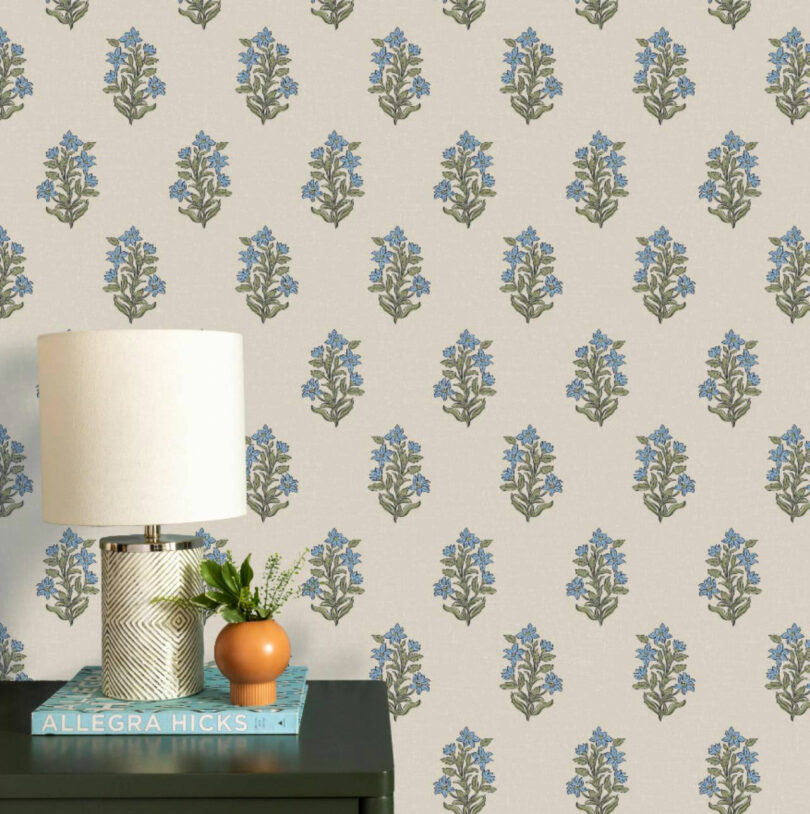 Table with a white lamp, two stacked books, and a small orange vase with greenery, set against Elizabeth Rees wallpaper featuring a repeating blue floral pattern.