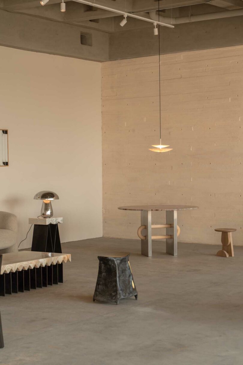 Minimalist room with modern furniture including a sofa, side table, lamps, and a round table with chairs. Concrete walls and floor, with a pendant light hanging from the ceiling