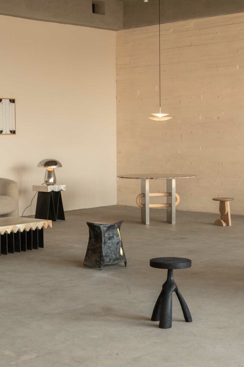 Minimalistic room with modern furniture, including a beige sofa, black and gray stools, a marble table, and pendant lighting, all set against light-toned walls and concrete floor