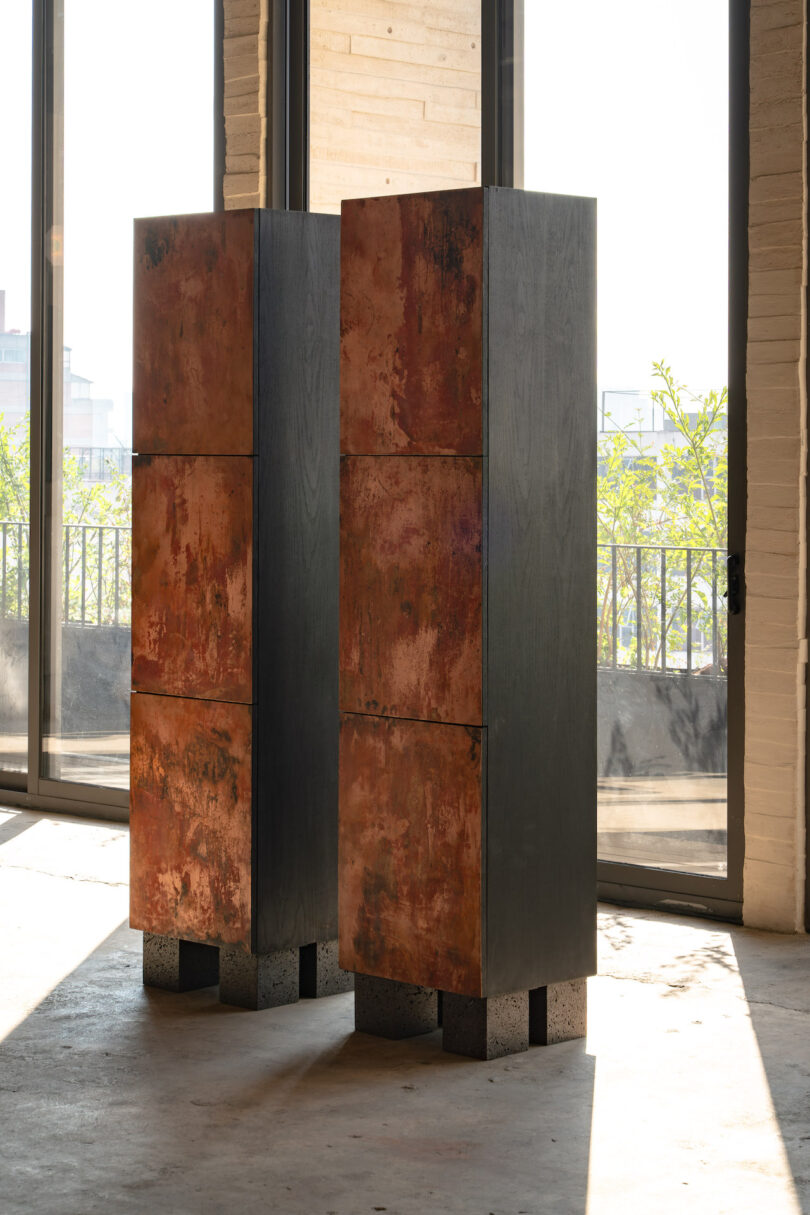 Two tall, rectangular sculptures with a rusty, textured surface and black bases, placed indoors near large windows with sunlight and cityscape in the background