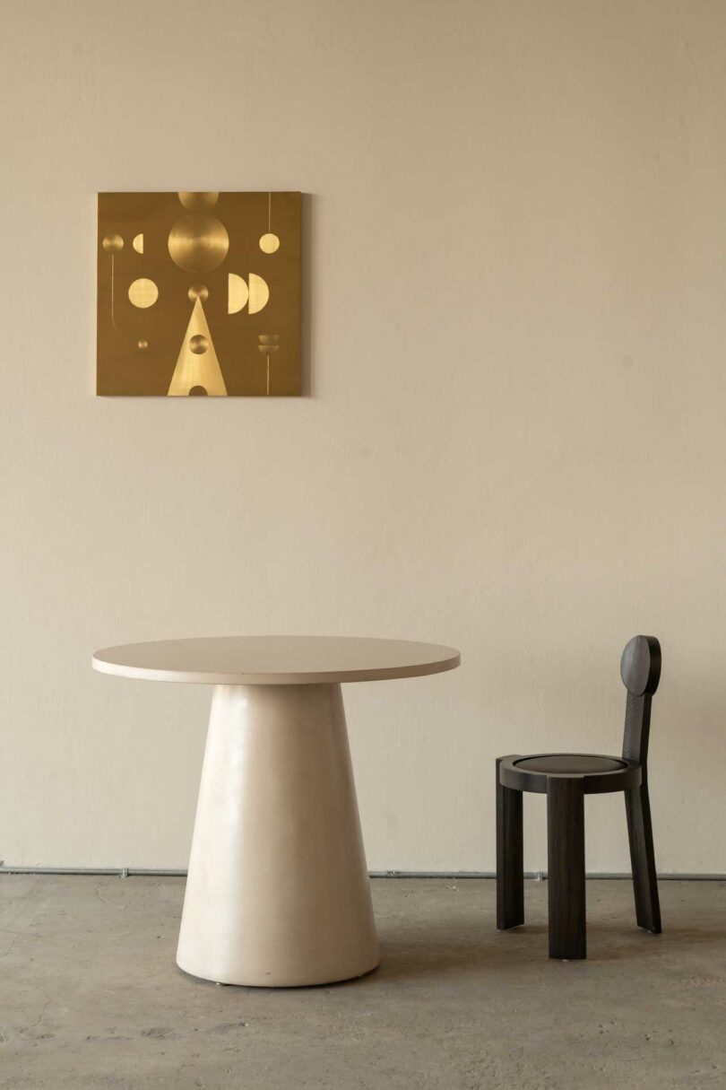 Minimalist room with a round table, a black chair, and a geometric wall art piece. The wall art features gold shapes on a mustard background. The floor is concrete and the wall is beige