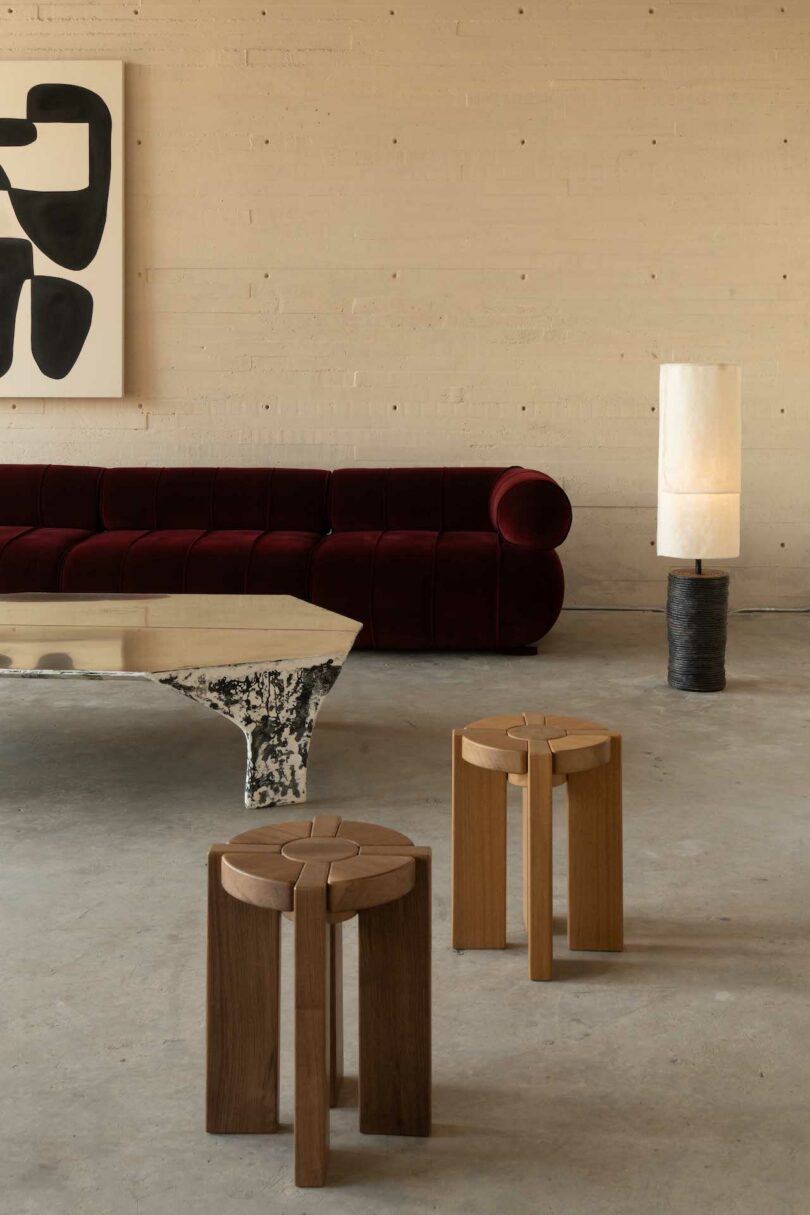 A modern living room with a maroon sofa, abstract wall art, two wooden stools, a metallic coffee table, and a floor lamp on a concrete floor against a light-colored wall