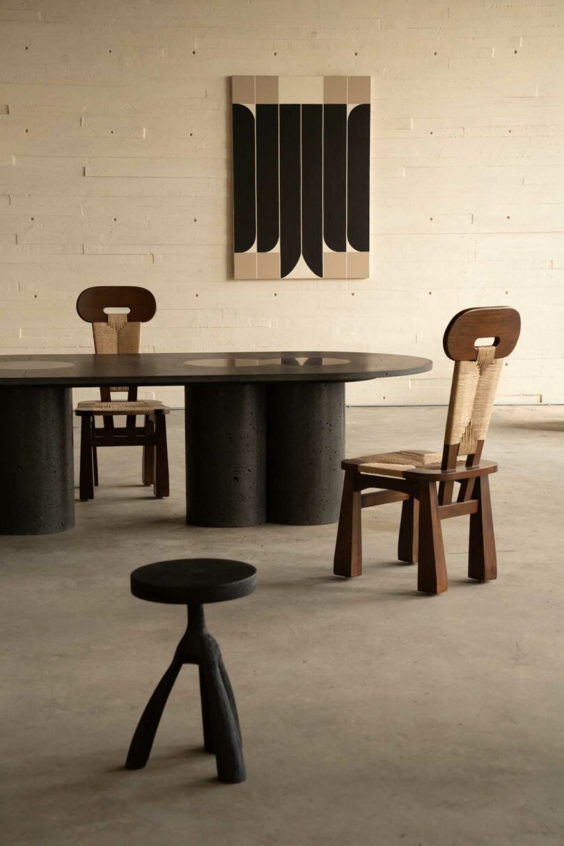 Minimalist interior with two wooden chairs, a round black table, and a small dark stool. A geometric artwork hangs on the beige wall