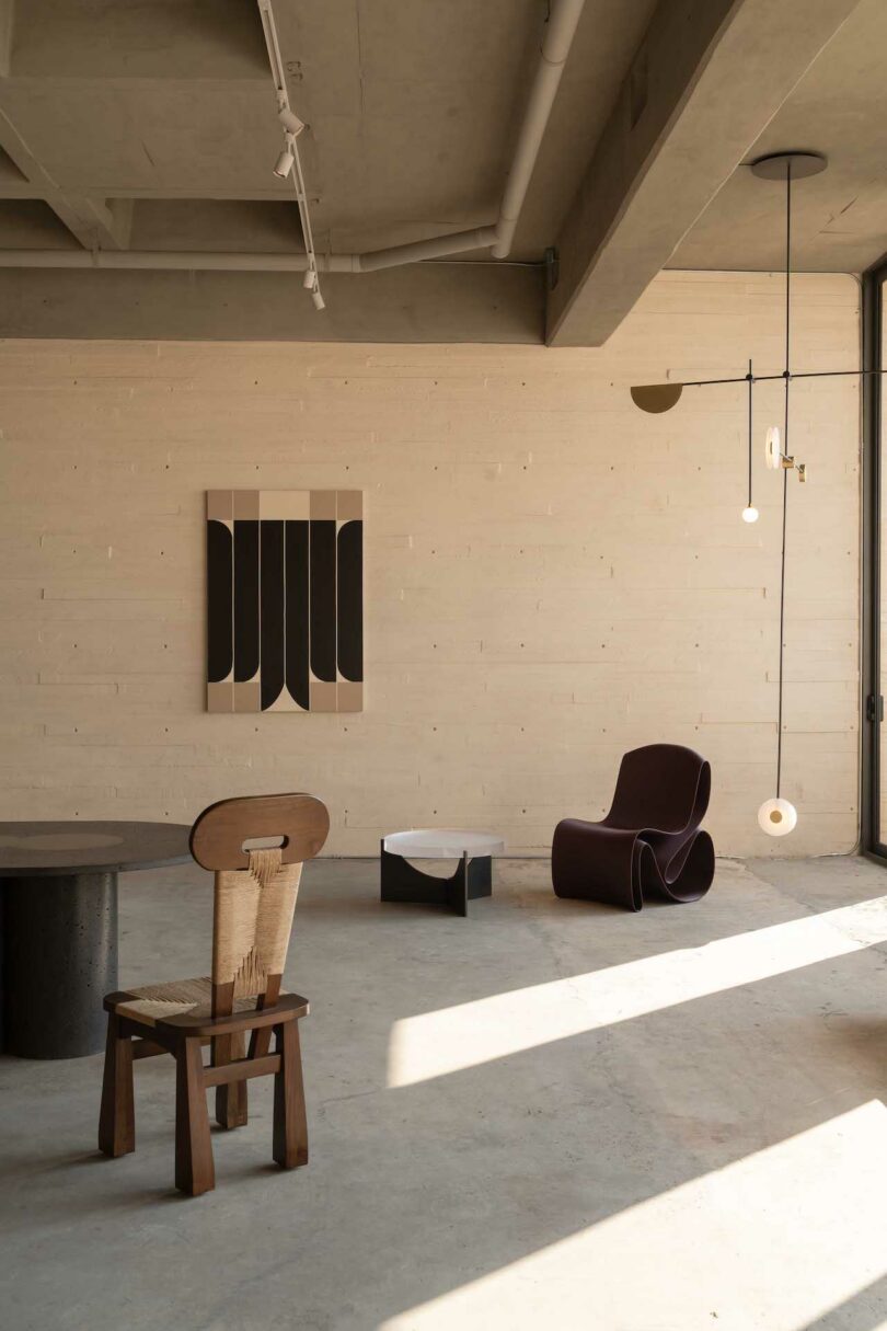 Minimalist room with a wooden chair, modern dark brown lounge chair, low black table, abstract wall art, and a hanging circular light fixture. Natural light casts shadows on the concrete floor