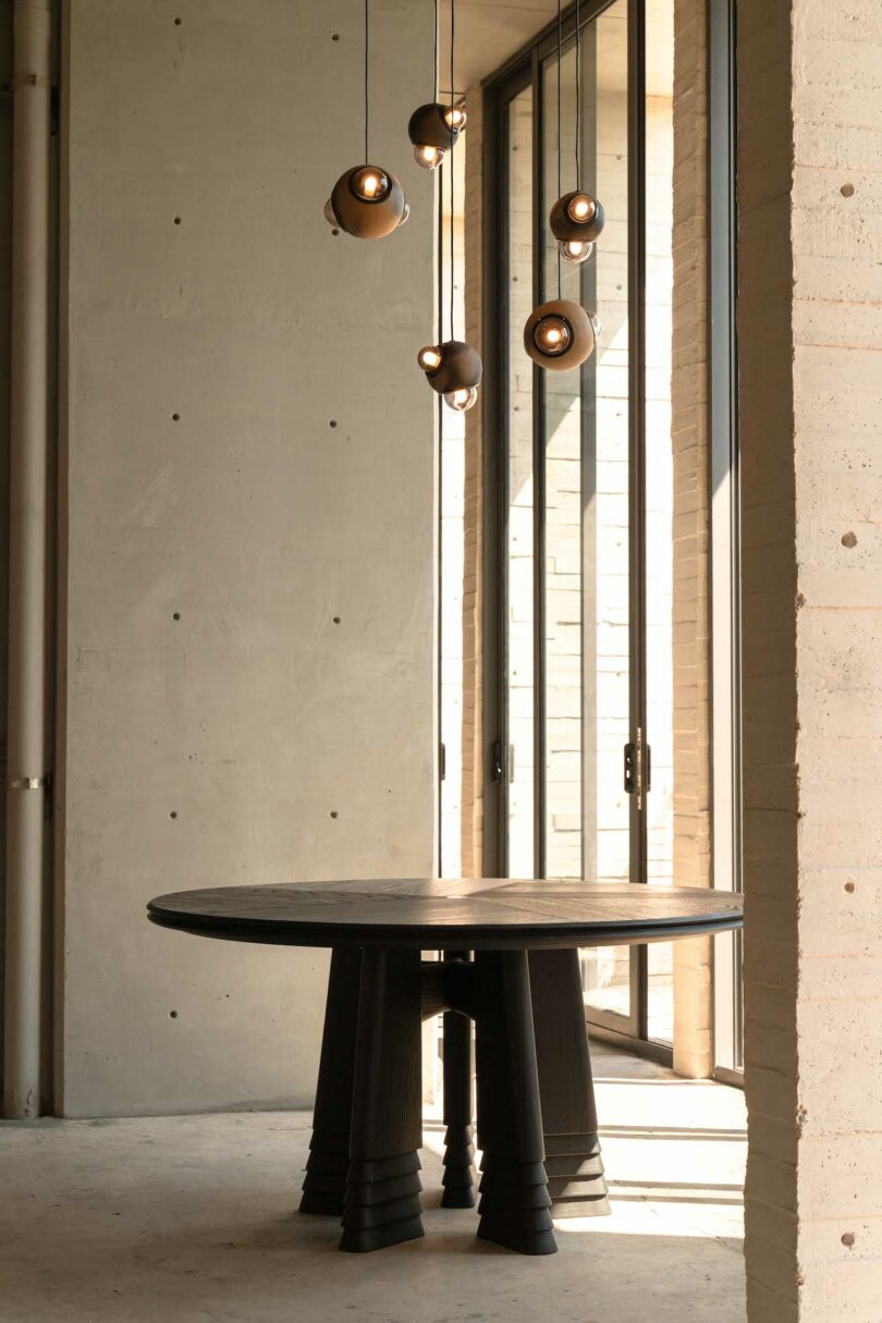 A round, dark wooden table beneath six hanging pendant lights near tall glass windows in an industrial-style space with concrete walls