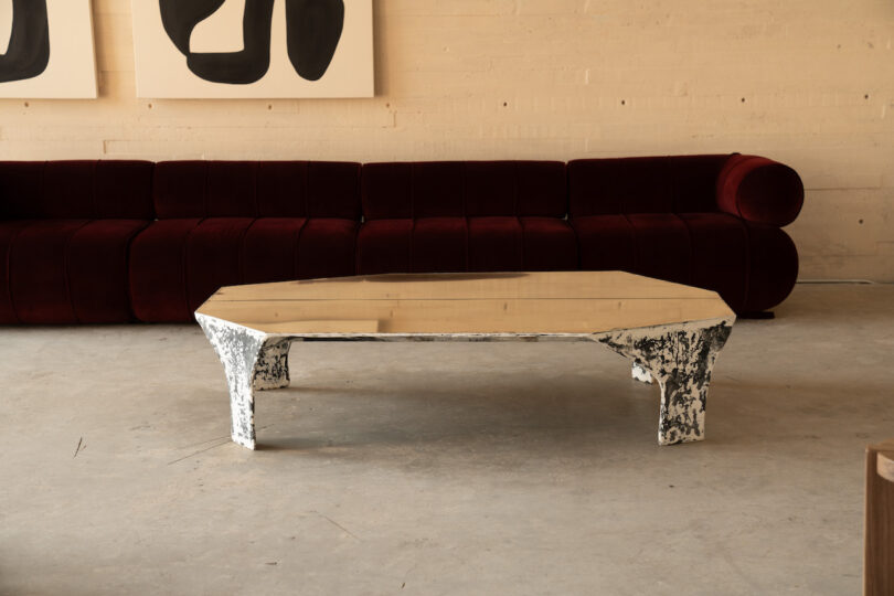 A hexagonal coffee table with textured white legs and a reflective top stands on a concrete floor. A velvet maroon sofa is in the background against a light-colored wall