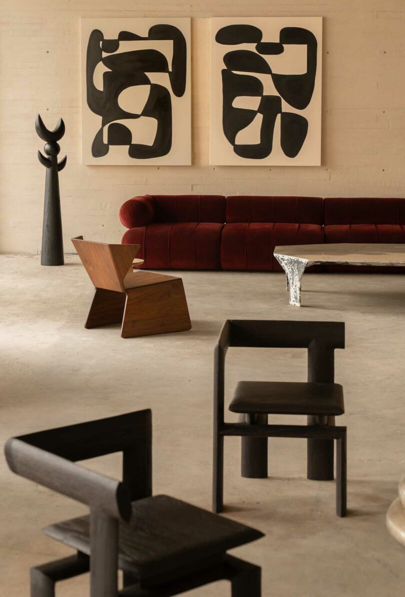 Modern living room with abstract black-and-white artwork, a red sofa, wooden and black chairs, and a tall sculptural piece. The room is minimalistic with concrete flooring