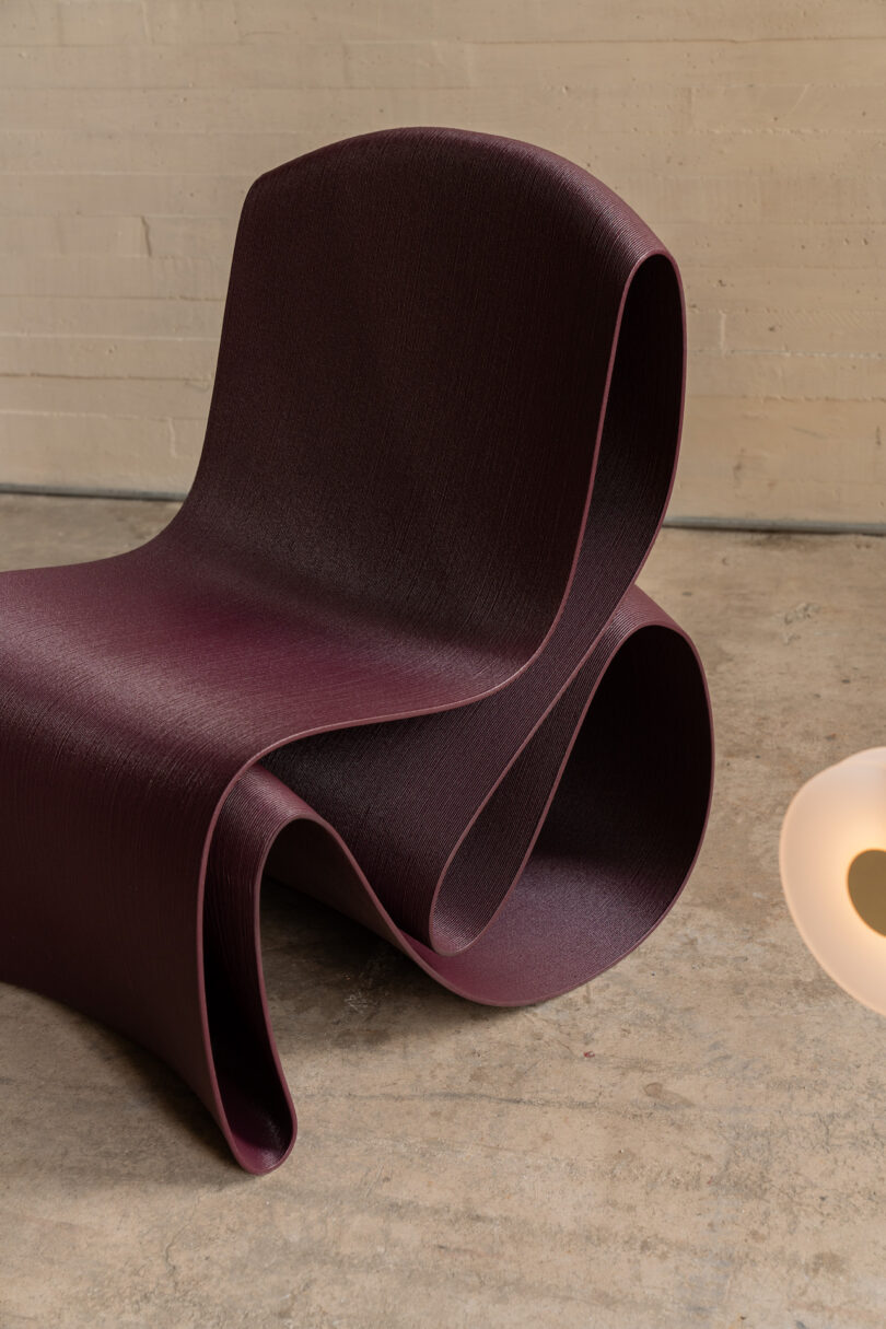 Modern, curvy dark purple chair on a concrete floor near a white, oval-shaped light fixture against a beige wall