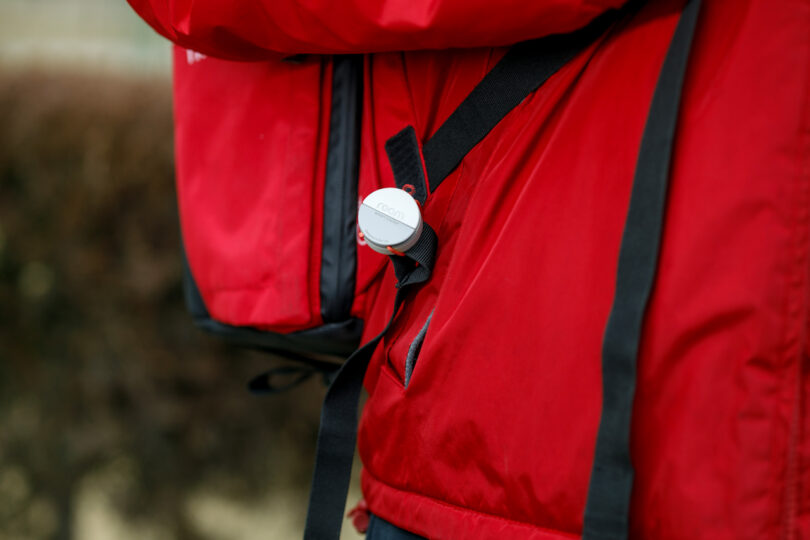 Close-up of a person wearing a red jacket and backpack, featuring a round button or pin attached to a black strap.