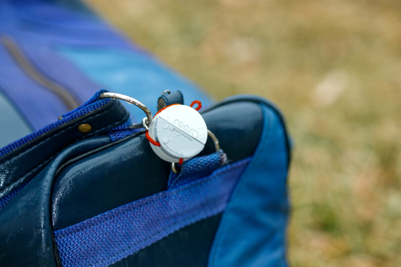 A blue bag with a white tracking device attached to the zipper pull via a metal ring. The device has a gray strap looped through it. The background is grassy.