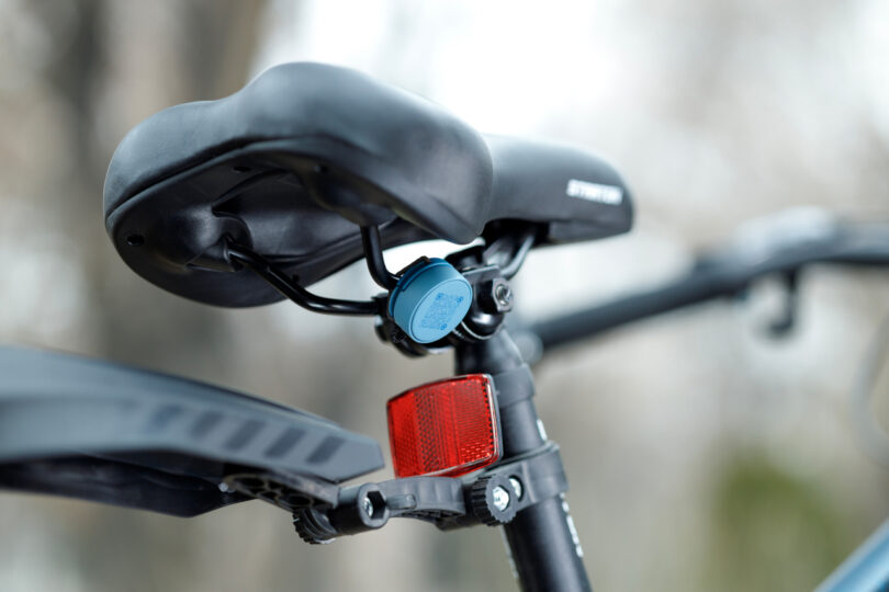 Close-up of a bicycle seat with a blue device attached underneath and a red reflector on the seat post.