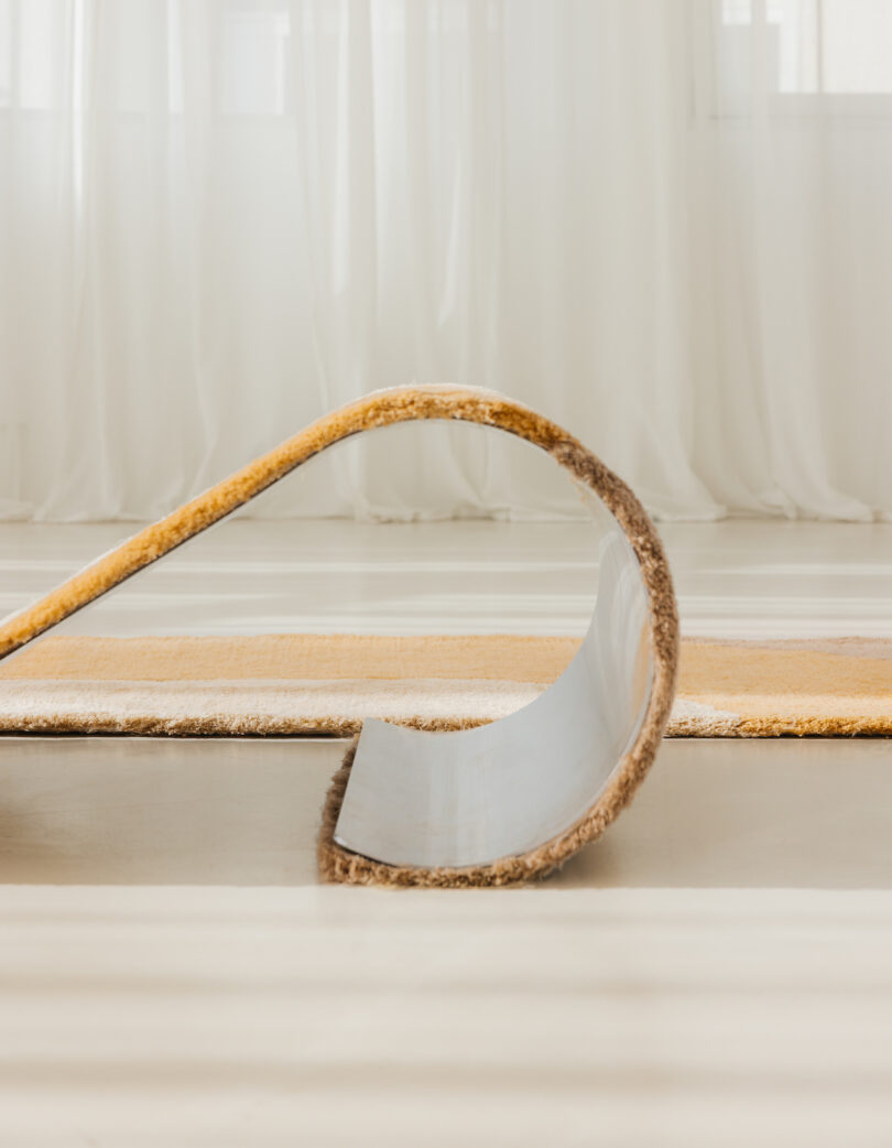 A chaise/rug hybrid, with its edge curling upwards, lies on a light-colored floor. Sheer curtains hang in the background.