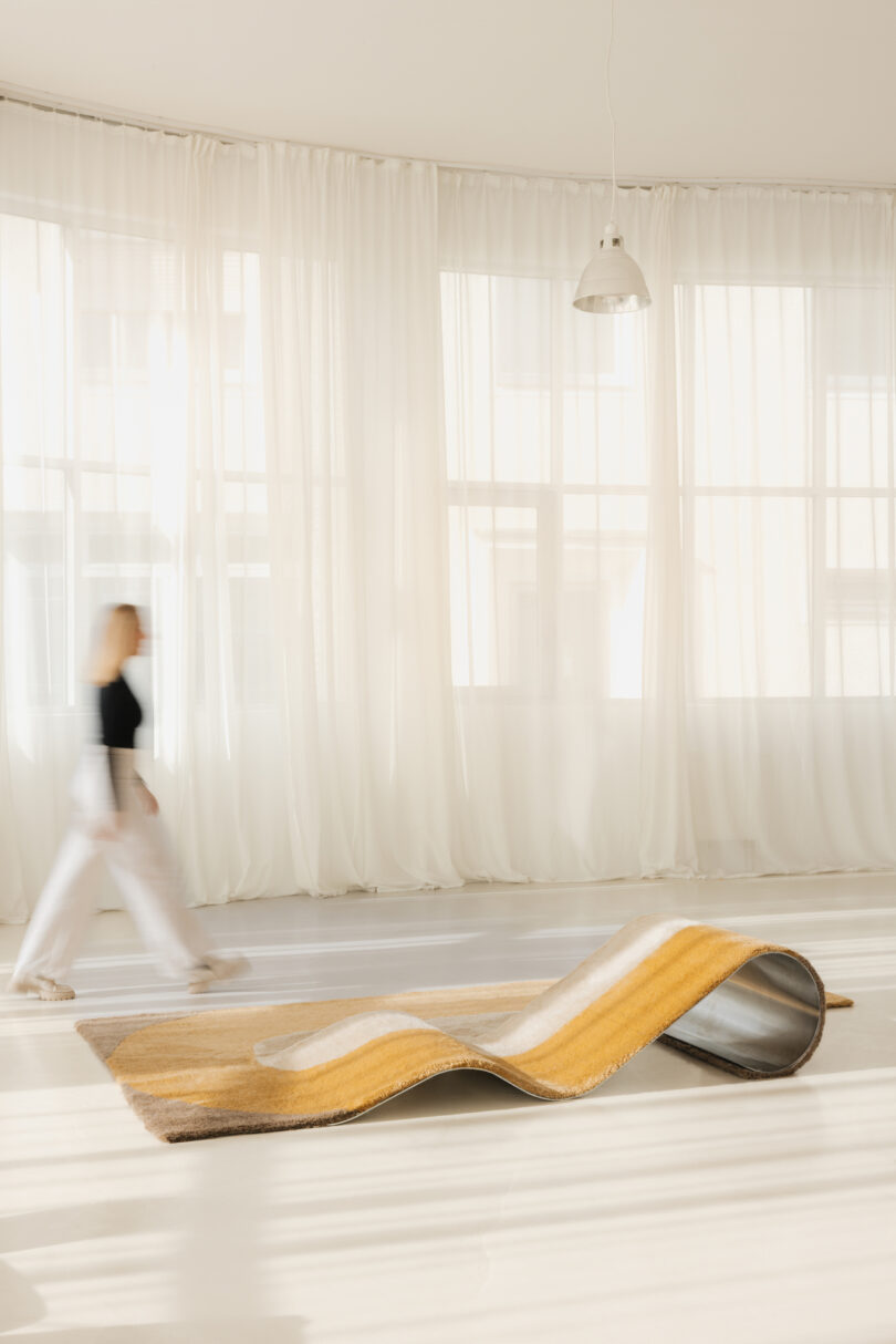 A person walks past a wavy, golden chaise/rug hybrid on a light floor with sheer curtains and large windows in the background.
