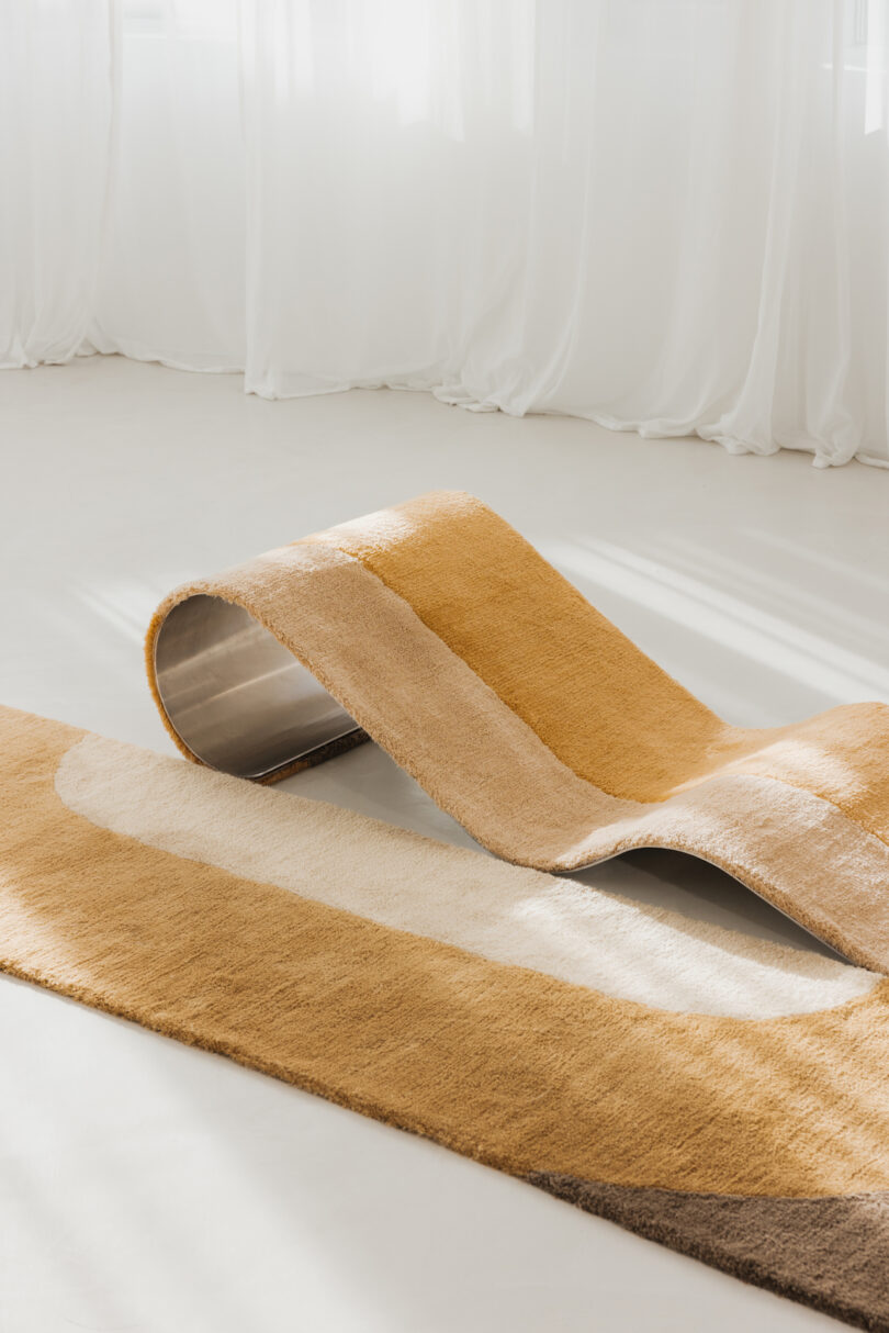 A curvy chaise/rug hybrid on a light floor with sheer white curtains in the background.