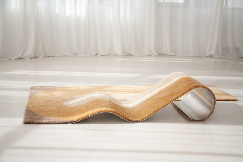 A curvy chaise/rug hybrid on a light floor with sheer white curtains in the background.