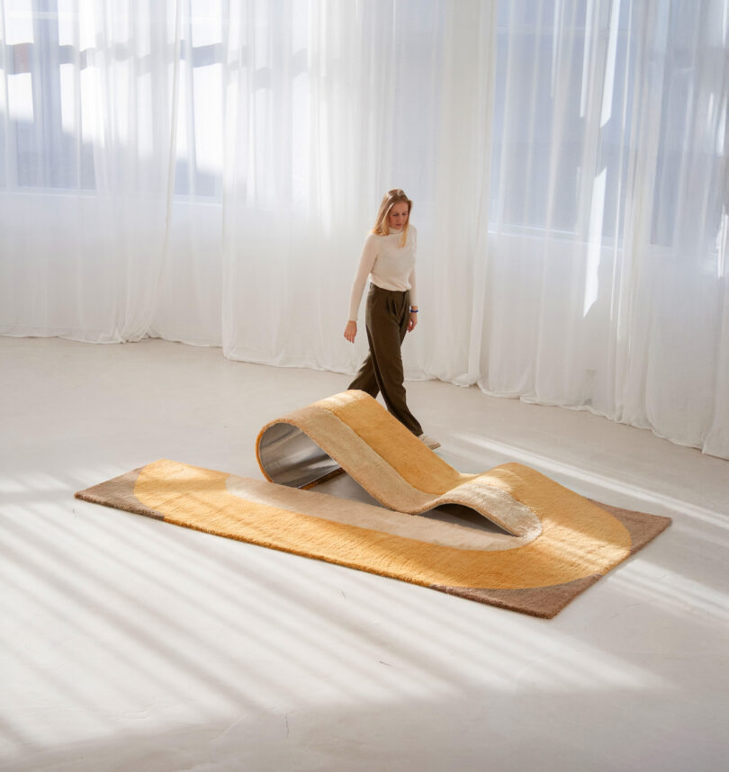 A person walks near a large, wavy, beige and tan chaise/rug hybrid in a sunlit room with white curtains.