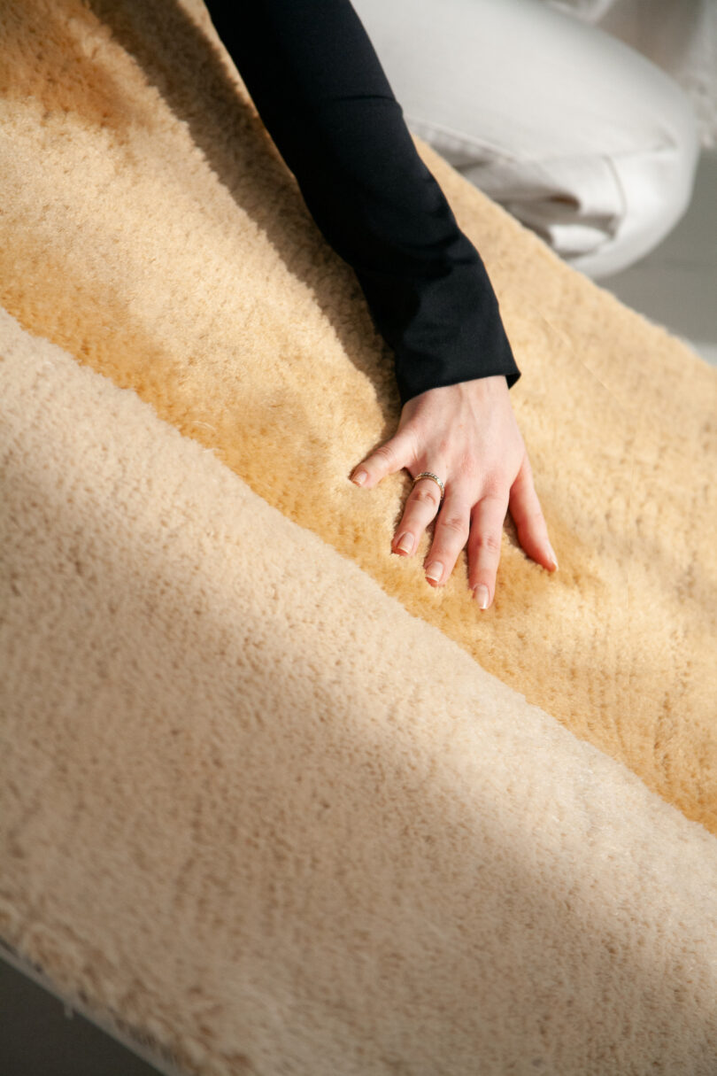 A person with a ring on their finger gently touches a soft, beige fur-like fabric surface