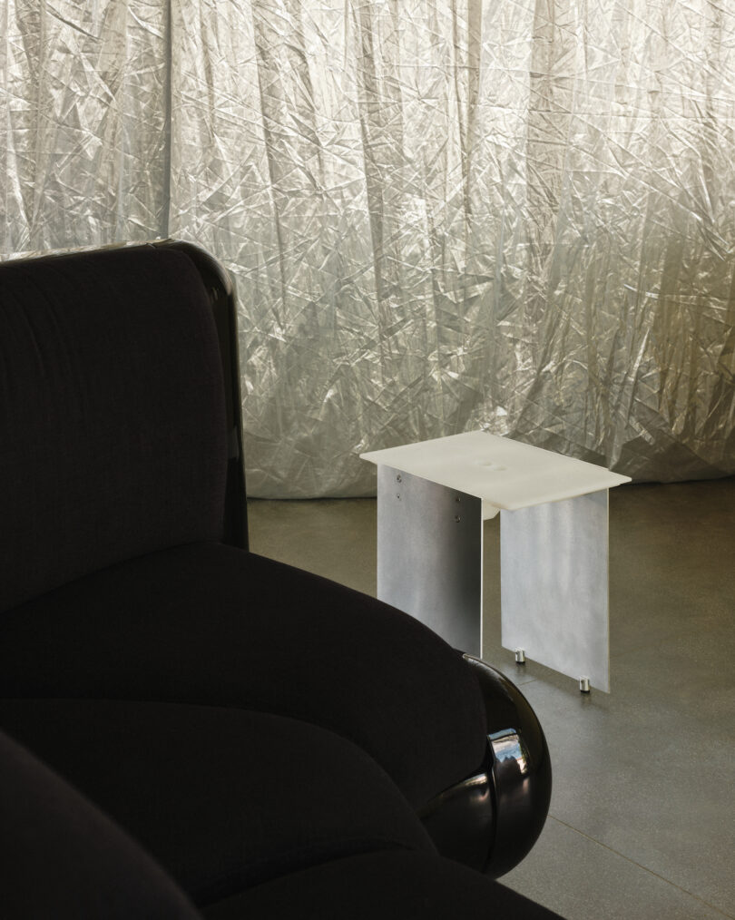 Black sofa and small white side table in front of a silver crinkled curtain on a concrete floor