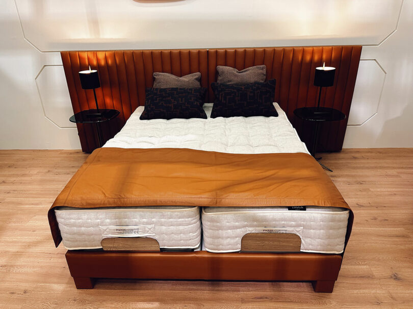 A neatly made bed with a padded brown headboard, four pillows, a brown throw blanket, and two round bedside tables with lamps, set on a wooden floor.