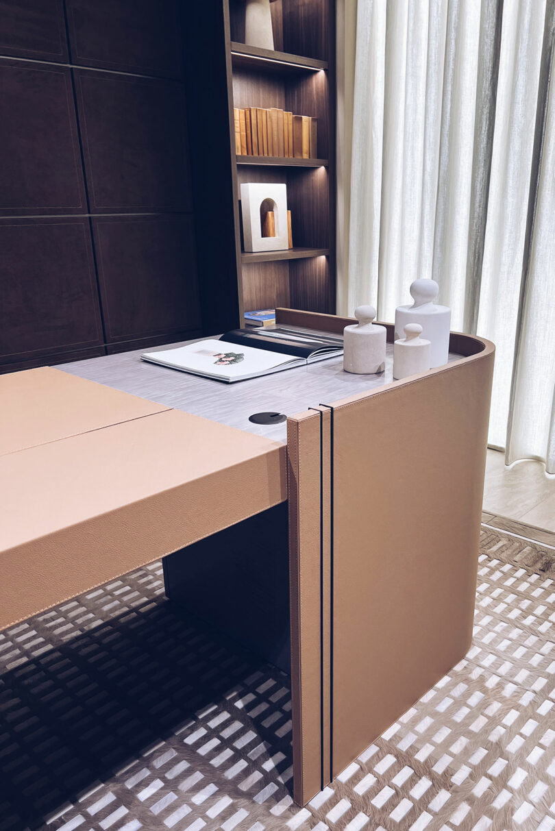 Modern office desk with a laptop, notebook, and decorative objects, positioned near a bookshelf and curtains. The area features a patterned rug and contemporary design elements.