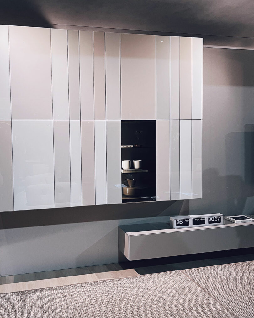 Modern wall-mounted white cabinets with vertical grooves, a central open shelf displaying dishes, and a floating drawer unit below; digital date and time display on the surface.