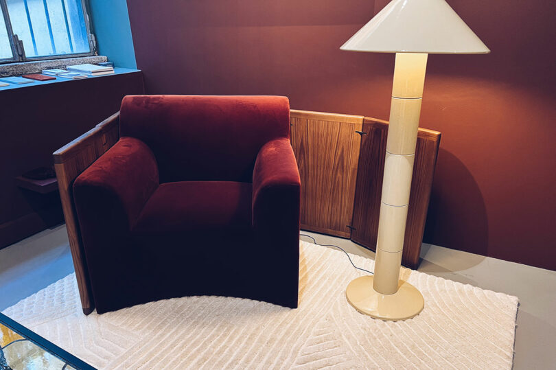 A burgundy velvet armchair sits next to a cream-colored floor lamp on a textured white rug, with wooden paneling and a window in the background.