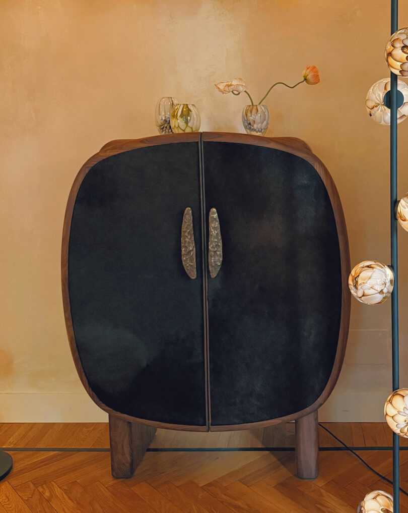 A dark wood cabinet with rounded edges and textured handles sits against a beige wall, with two small vases holding flowers on top and a lit string of globes to the right.