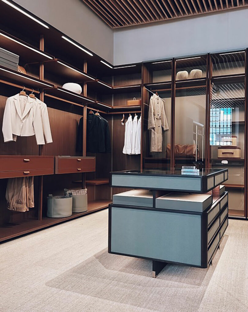 Spacious modern walk-in closet with wooden shelves, drawers, hanging clothes, and a central island—reminiscent of a Salone showroom—beautifully lit by natural and artificial light.