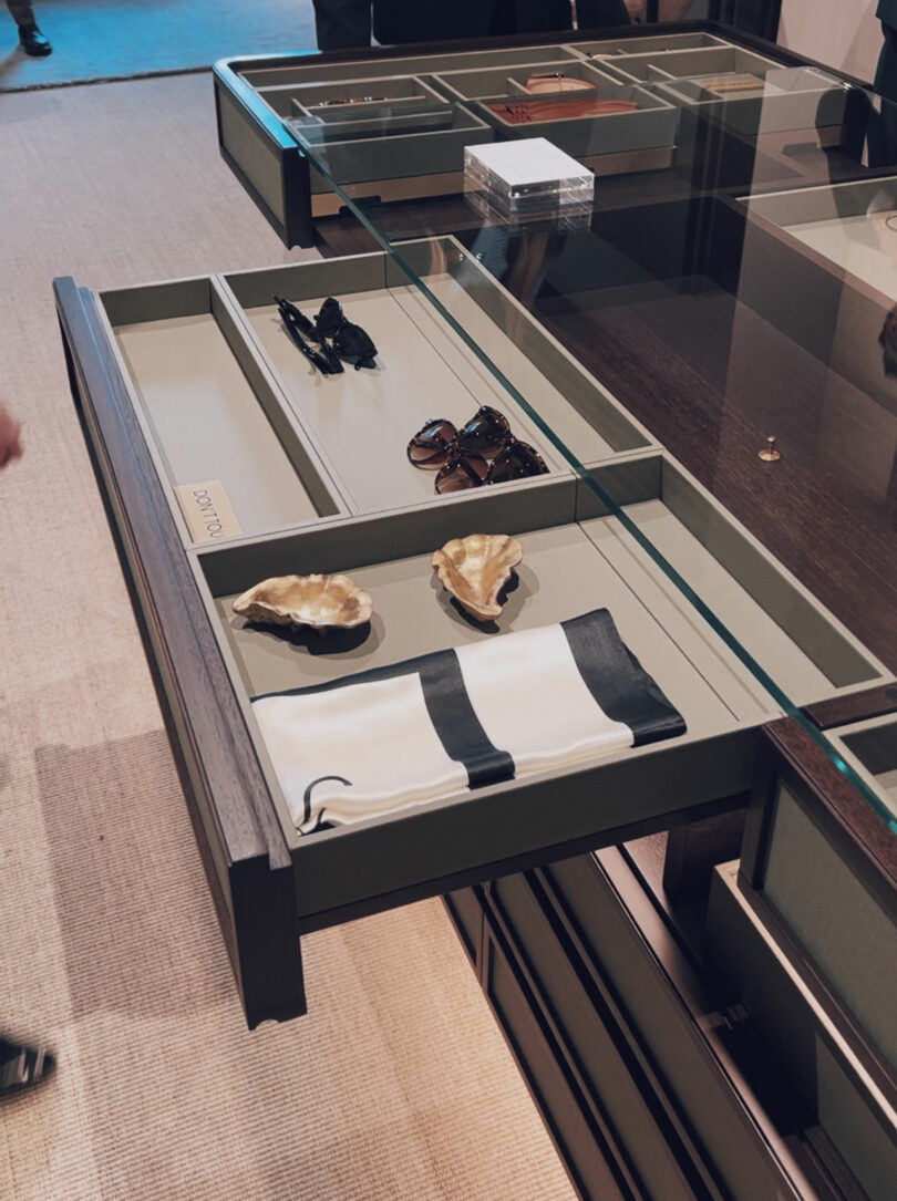 An open display drawer showcases sunglasses, gloves, two decorative shells, and a folded black-and-white cloth under a glass counter.