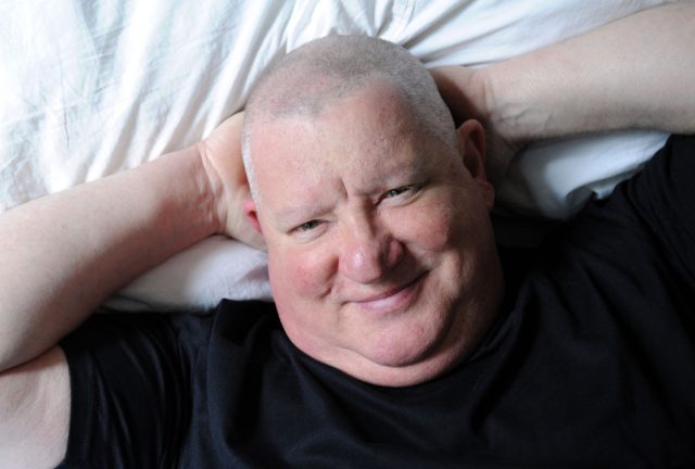 A man lies back on a bed with his hands behind his head.