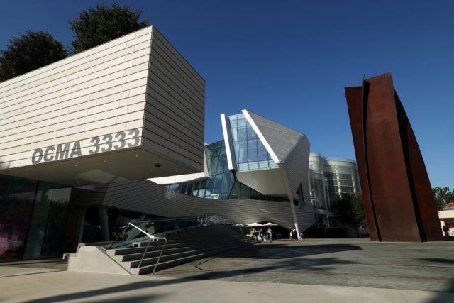 Exterior of a newly built museum showing three distinct structures. At left is rectangular shape made of light wood that juts over a small staircase. At center is a silver-ish building with angles and glass. At right is a Richard Serra sculpture made of brown steel.