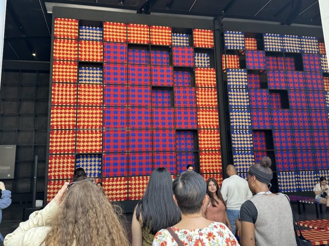 People gaze at a 25-foot tall installation made of woven squares in red, blue, and gold.