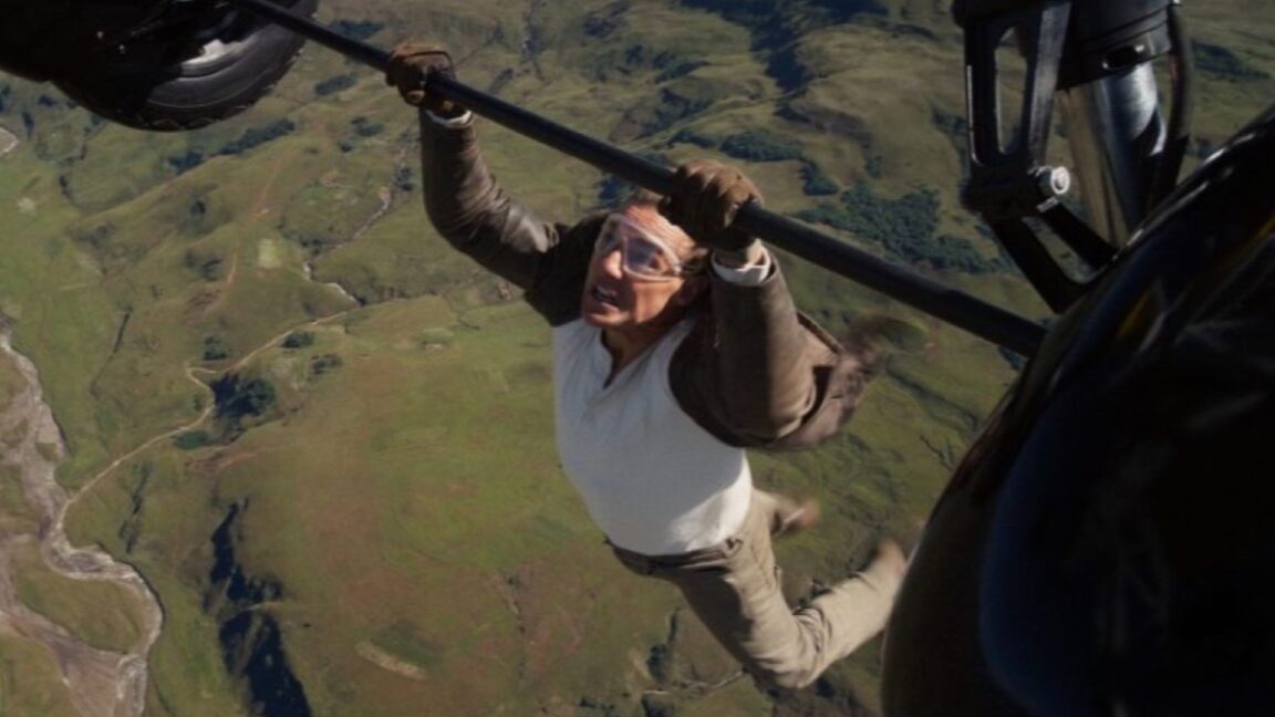 tom cruise hanging off landing gear of a yellow plane mid flight.