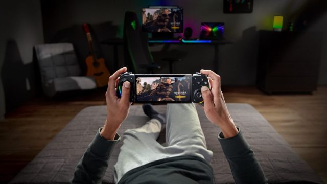 Razer-built-a-game-streaming-app-on-top-of-Moonlight-and.jpeg A person sitting very laid back on a couch holds up a phone with The Witcher III on it, with Razer Kishi controller around it, and a gaming PC showing the same scene in the distance.
