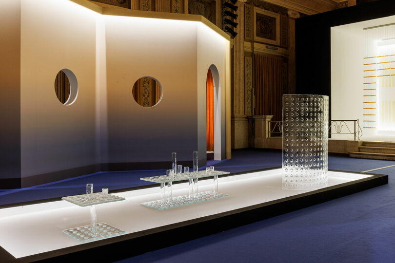 A modern exhibition space by POETICA Wonderglass features glass tables and cylinders on a raised platform, set against a curved wall with circular cutouts in the background.