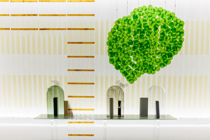 A modern interior wall with gold and white stripes, green circular wall art, and a glass shelf holding minimalist vases and geometric objects.