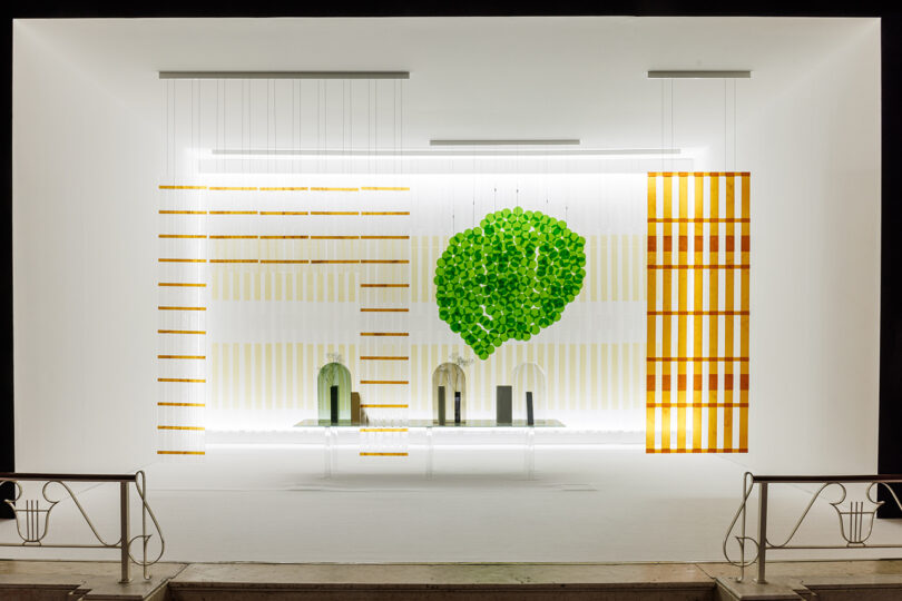 A modern art installation in a grand hall features a transparent cylindrical sculpture with circular cutouts and a stage backdrop with geometric patterns and a green tree design.